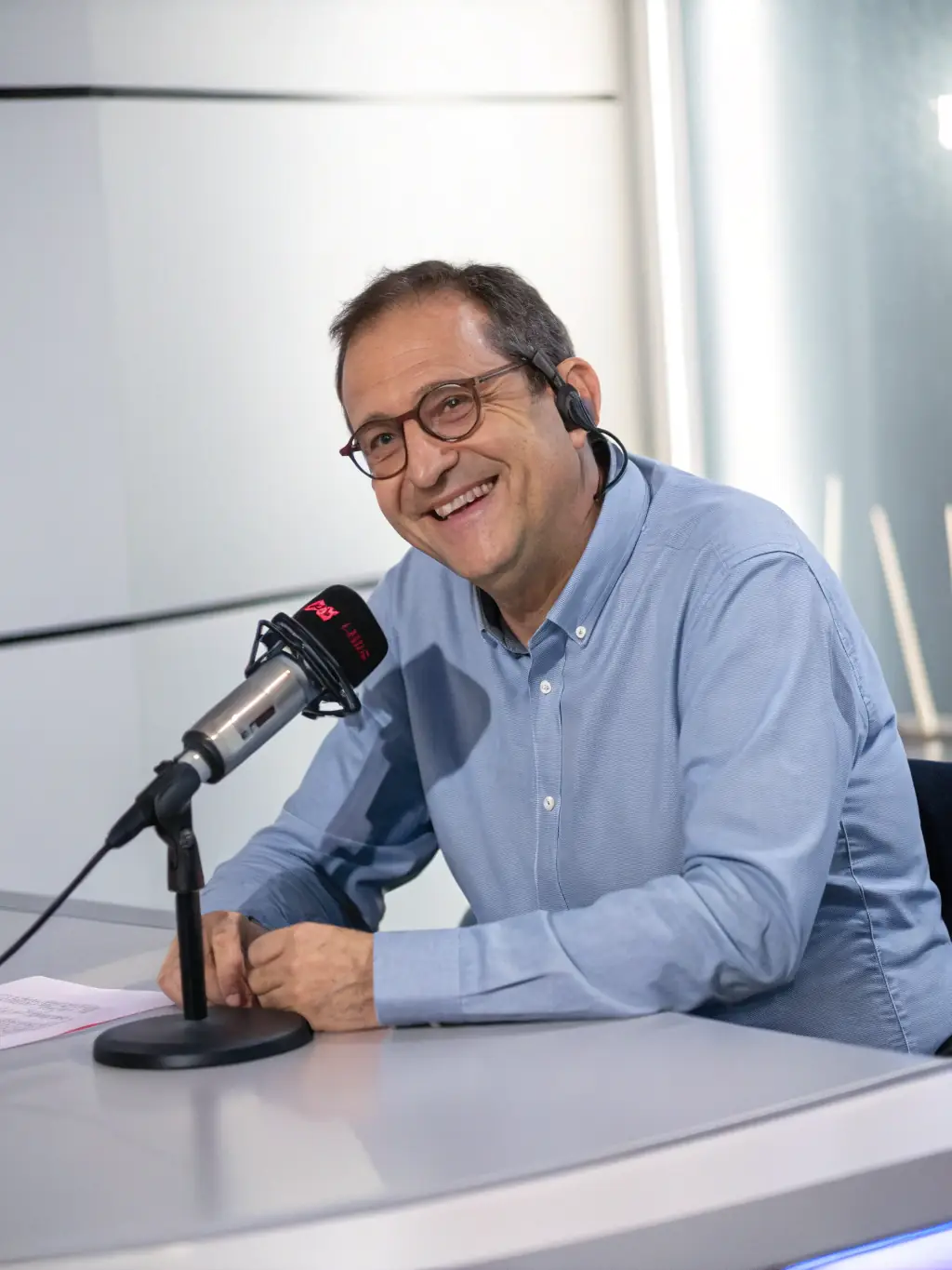 A professional headshot of a male radio host in his 50s, smiling confidently in a radio studio with microphones and sound equipment visible in the background, representing the host of 'Jeffco Morning Drive'.
